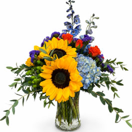 Bouquet of sunflowers, blue hydrangea, orange roses, and purple flowers in a glass vase