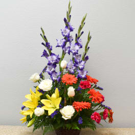 Tall mixed flower arrangement with purple spikes, yellow lilies, and orange daisies in a basket