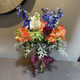 Colorful bouquet of orange, blue, and white flowers in a glass vase with a purple ribbon