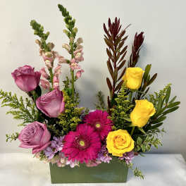Rectangular arrangement of lavender roses, bright pink gerbera daisies, and yellow roses in a green container