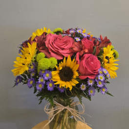 Bouquet of pink roses, sunflowers, and purple daisies in a glass vase