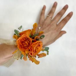 Wrist corsage with bright orange roses and accent blooms on a gold ribbon, worn on a hand.