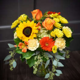 Mixed bouquet with sunflowers, orange roses, gerbera daisies, and yellow mums in a glass vase