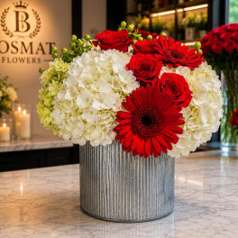 Red roses and gerbera daisies arranged with white hydrangeas in a ribbed metal vase.