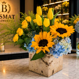 Yellow tulips, sunflowers, roses, and blue hydrangeas in a square vase