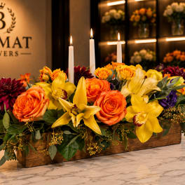 Long floral centerpiece with orange roses, yellow lilies, and lit candles in a wooden box.