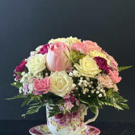 Compact arrangement of pink and white roses and mixed blooms in a floral teacup with matching saucer.