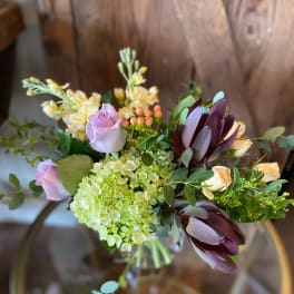 Arrangement with green hydrangea, lavender roses, cream blooms and dark burgundy protea in a glass vase