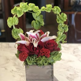 Red roses and pink lilies in a gray square vase with a green floral arch
