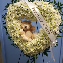 Heart-shaped white flower wreath with a teddy bear and ribbon on a stand