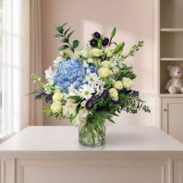 Mixed bouquet of blue hydrangeas, white daisies, and pale roses in a glass vase