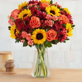 Bouquet of sunflowers, orange roses, and red mums in a clear glass vase