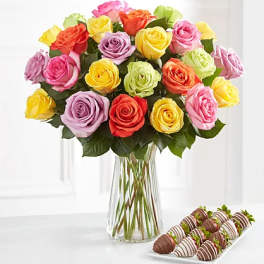 Colorful rose bouquet in a clear glass vase with chocolate-covered strawberries beside it