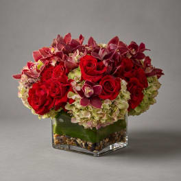Red roses and purple orchids arranged in a square glass vase with pebbles.
