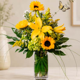 Yellow lilies and sunflowers in a clear glass vase with a butterfly pick