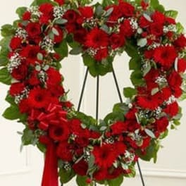 Heart-shaped standing wreath of red flowers on a metal easel with a red ribbon bow.