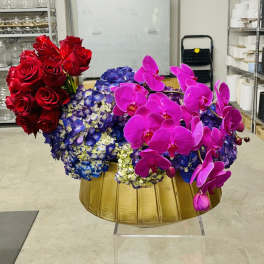 Red roses, blue hydrangeas, and pink orchids arranged in a gold bowl