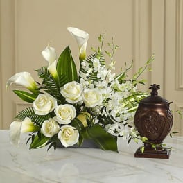 White roses and calla lilies arranged with greenery beside a dark urn.