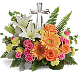 Low arrangement of white lilies, orange gerbera daisies, roses and pink spray roses around a silver cross