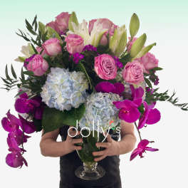 Tall arrangement of pink roses, white lilies, blue hydrangeas, and fuchsia orchids in a clear glass vase