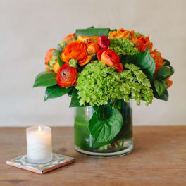 Orange and green floral arrangement in a glass vase beside a lit candle
