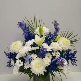 Tall arrangement of white lilies, roses, mums, and blue flowers in a green glass vase