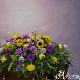 Colorful casket spray of yellow sunflowers, purple mums, and lavender and yellow roses on a wooden casket.