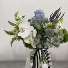 Low glass vase arrangement with white orchids, blue flowers, and green accents tied with a striped ribbon.