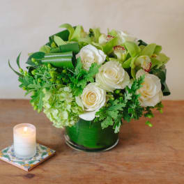 White roses and green orchids in a green glass vase beside a lit candle