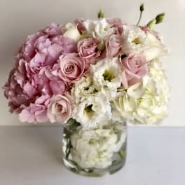 Low round arrangement of pink hydrangeas, pale pink roses, and white flowers in a clear glass vase.