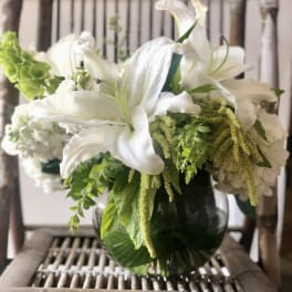 White lilies and mixed white and green flowers in a round glass vase.