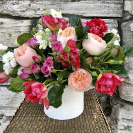 Low arrangement of pink and peach roses, tulips, and white stock in a white ceramic vase