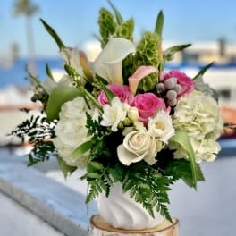 White and pink roses, calla lilies, and hydrangeas arranged in a white vase on a wooden base outdoors