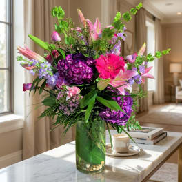 Tall bouquet of pink lilies, purple hydrangeas, and a pink gerbera daisy in a glass vase.