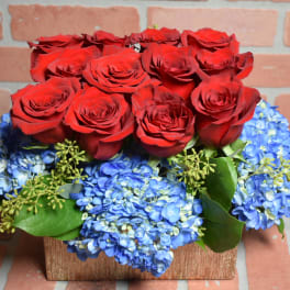 Red roses arranged above blue hydrangeas in a rectangular wooden container