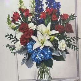 Bouquet of red roses, white lilies, blue flowers, and greenery in a clear vase