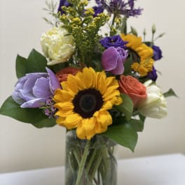 Mixed bouquet with a sunflower, roses, and purple blooms in a glass vase