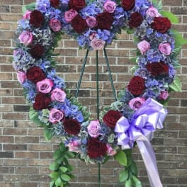 Heart-shaped standing wreath of red and pink roses with purple hydrangeas and a lavender bow on an easel.
