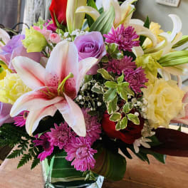 Mixed arrangement of pink lilies, purple roses, red roses, and yellow blooms in a square glass vase