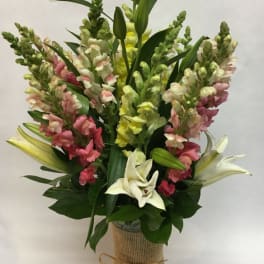 Tall bouquet of pink, yellow, and white snapdragons with white lilies in a burlap-wrapped vase