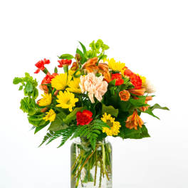Bright mixed bouquet of yellow daisies, orange alstroemeria, and red and peach carnations in a clear glass jar.