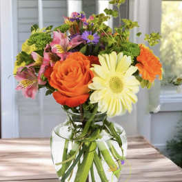 Mixed bouquet of orange, yellow, pink, and purple flowers in a clear glass vase.