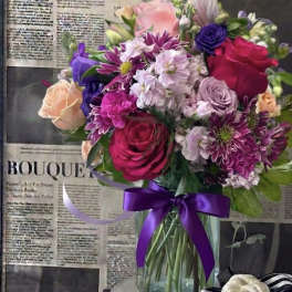 Mixed bouquet of pink, purple, and peach flowers in a glass vase with a purple ribbon