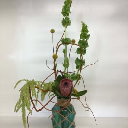 Tall floral arrangement in a teal glass vase with a pink protea and green blooms