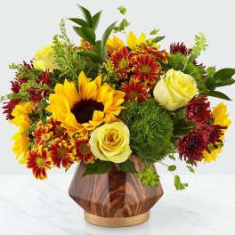 Short arrangement of sunflowers, yellow roses, and red chrysanthemums in a faceted wood-look vase