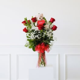 Red roses in a glass vase with a teddy bear and heart topper