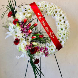 Heart-shaped funeral wreath with white daisies, lilies, and red ribbon on a stand