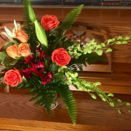 Mixed arrangement of orange roses, white lilies, red blooms, and green orchids in a clear glass vase
