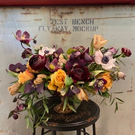 Mixed bouquet of purple, peach, and yellow flowers in a gold vase