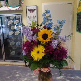 Mixed bouquet with sunflowers, purple daisies, and blue delphinium in a glass vase